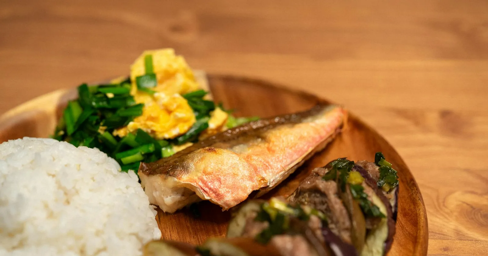 A wooden plate featuring white rice, grilled fish, sauteed vegetables, and scrambled eggs, showcasing Asian cuisine.