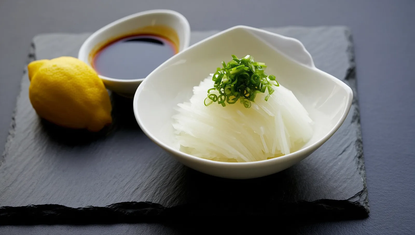 A white ceramic dish of finely grated daikon topped with sliced scallion, next to a saucer of dark ponzu and a lemon on slate.