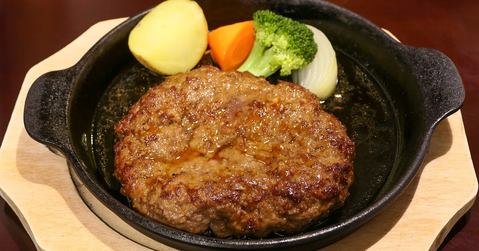 A hamburg steak sizzling on a black cast iron plate with boiled potato, carrot, broccoli and onion.
