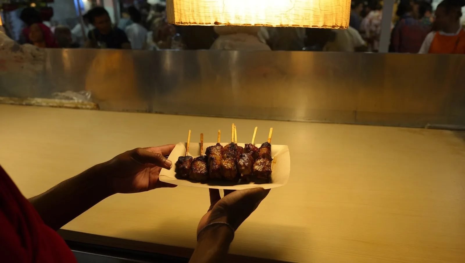 Two hands holding a small paper tray of glazed grilled skewers at a lantern-lit night market counter.