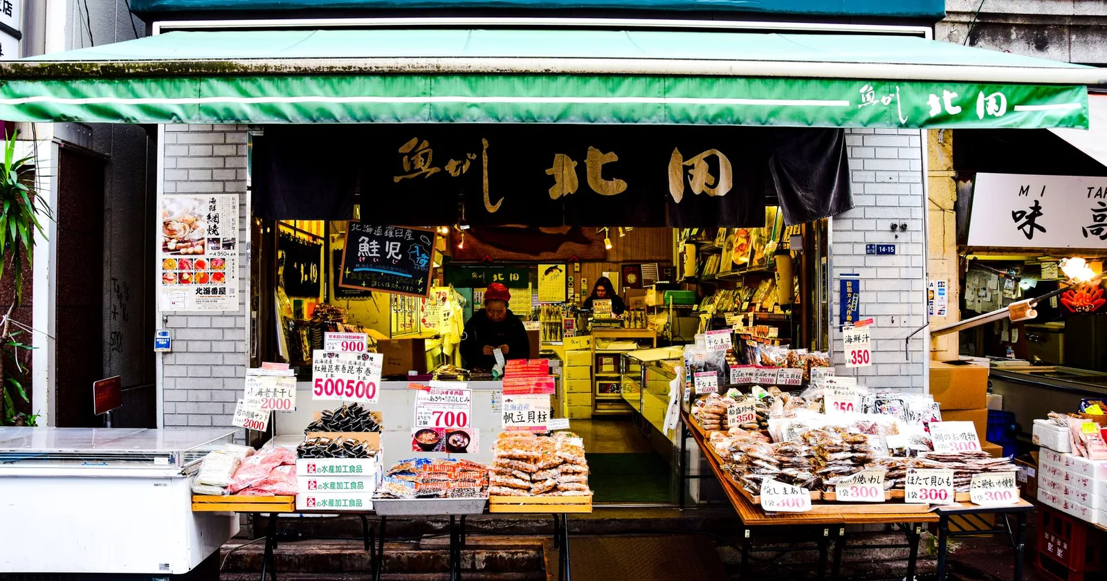 A Japanese street market food stall with hand-written price cards and trays of dried seafood and produce laid out on tables.