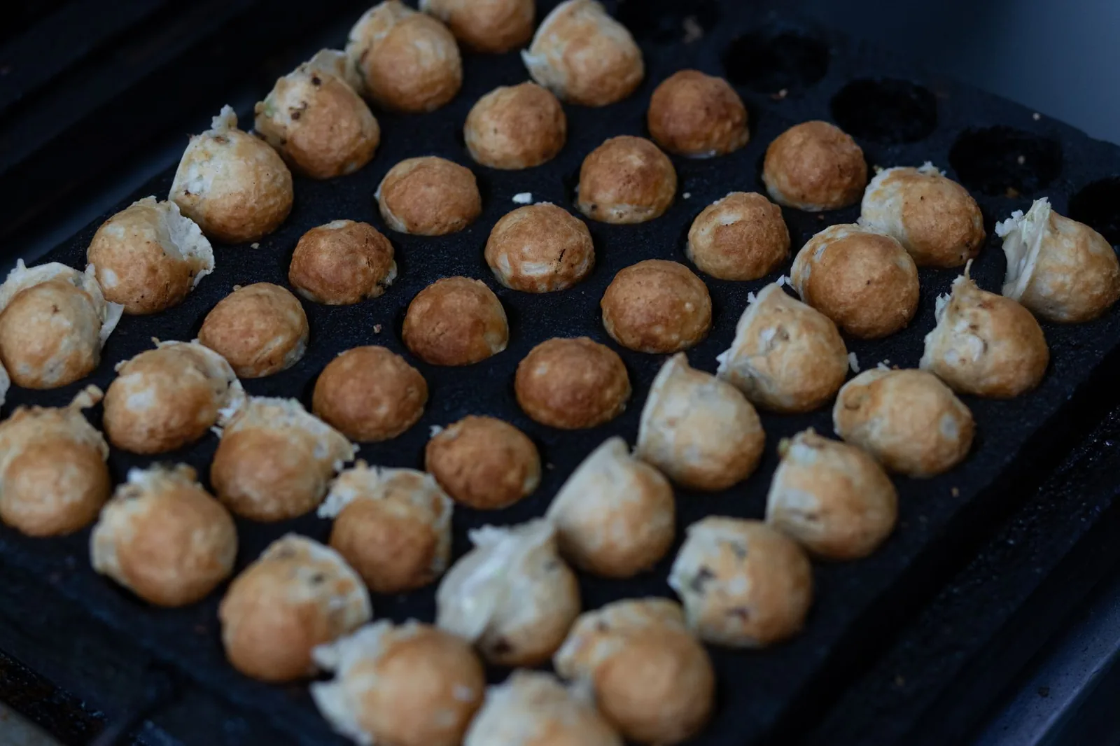 Takoyaki batter balls browning in the half-round molds of a cast-iron takoyaki pan.