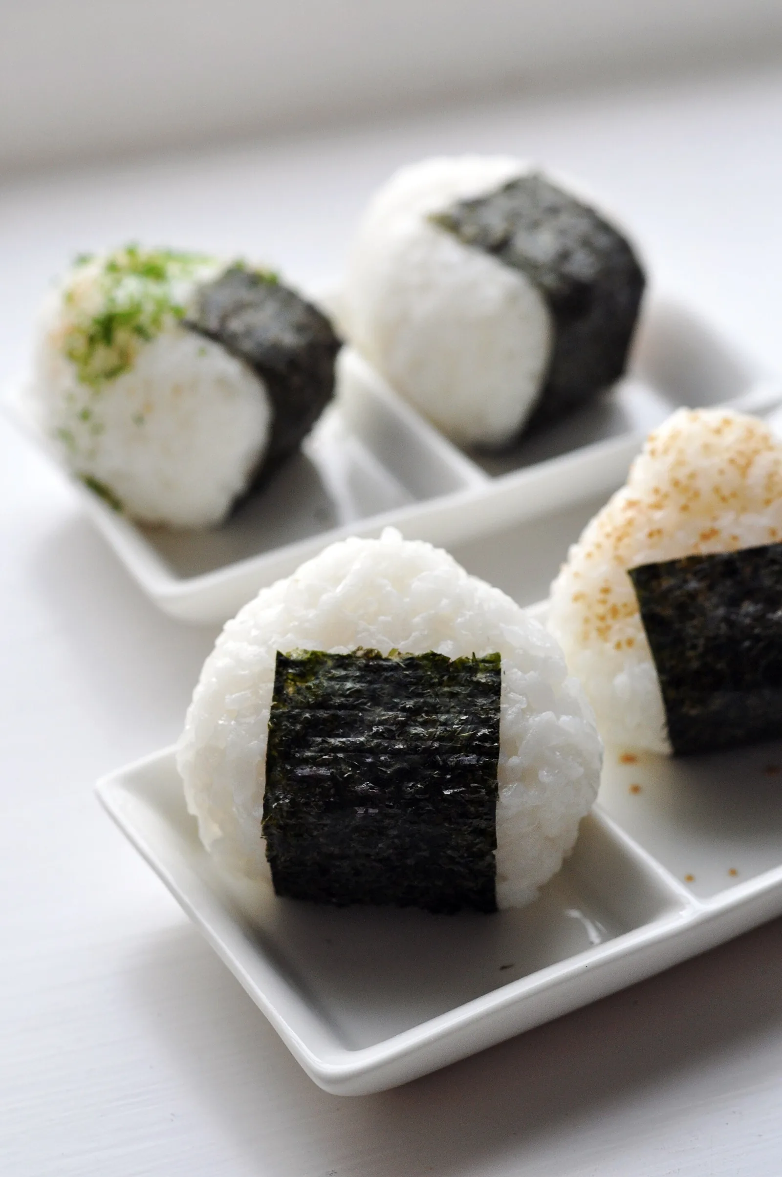 Four homemade onigiri on white square plates, each with a band of dark nori seaweed, one topped with green herbs and one with sesame.