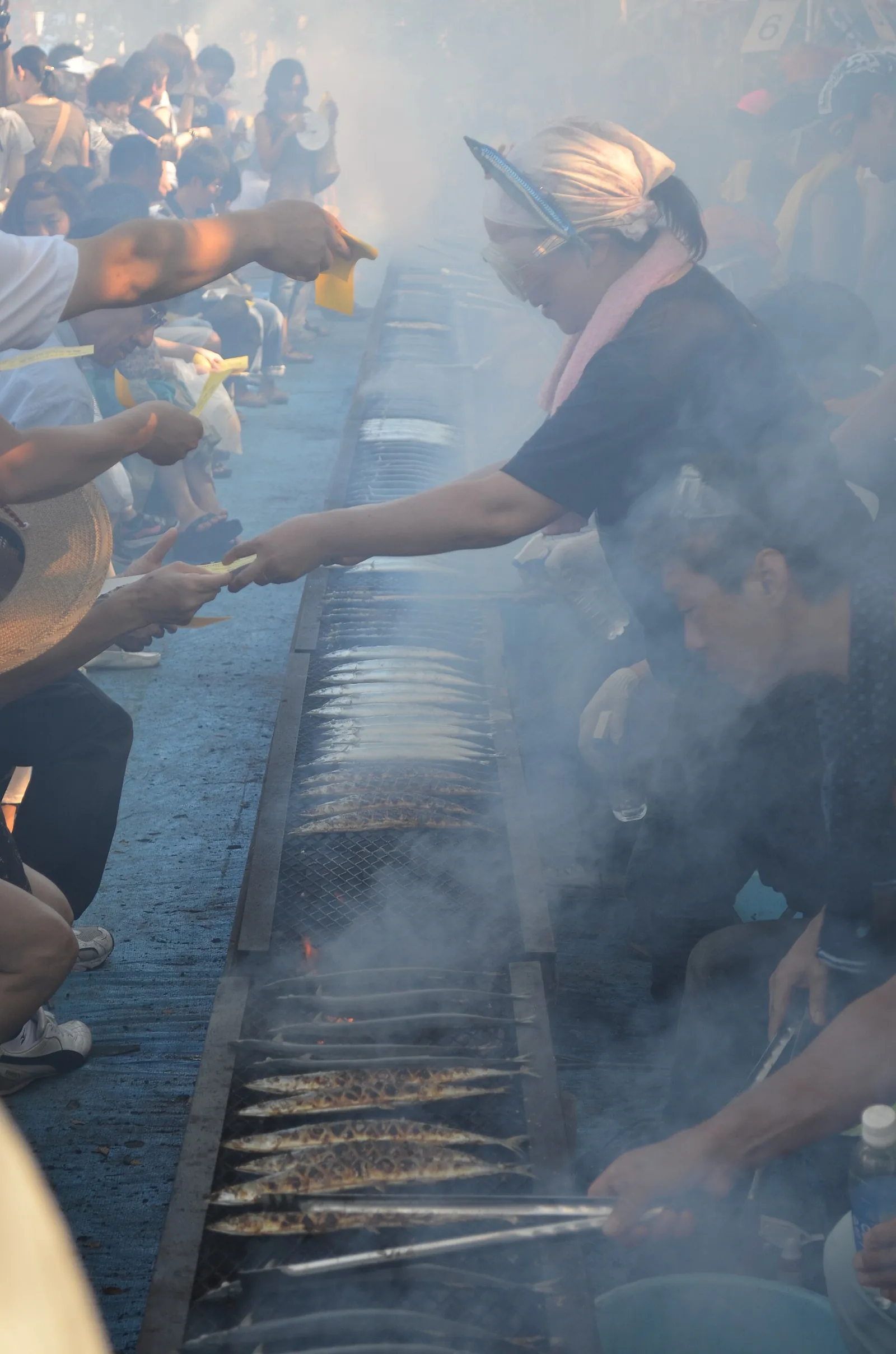 Rows of saury grilling over a long charcoal trench in heavy smoke