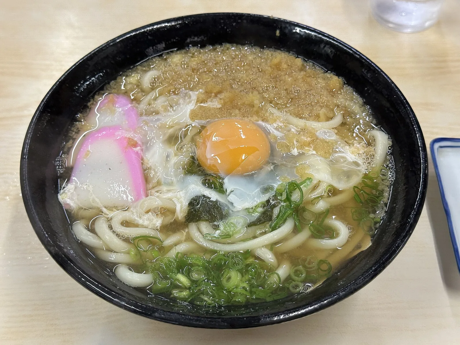 A bowl of tsukimi udon with an intact raw egg yolk, tempura crumbs, nori, green onion and pink kamaboko.